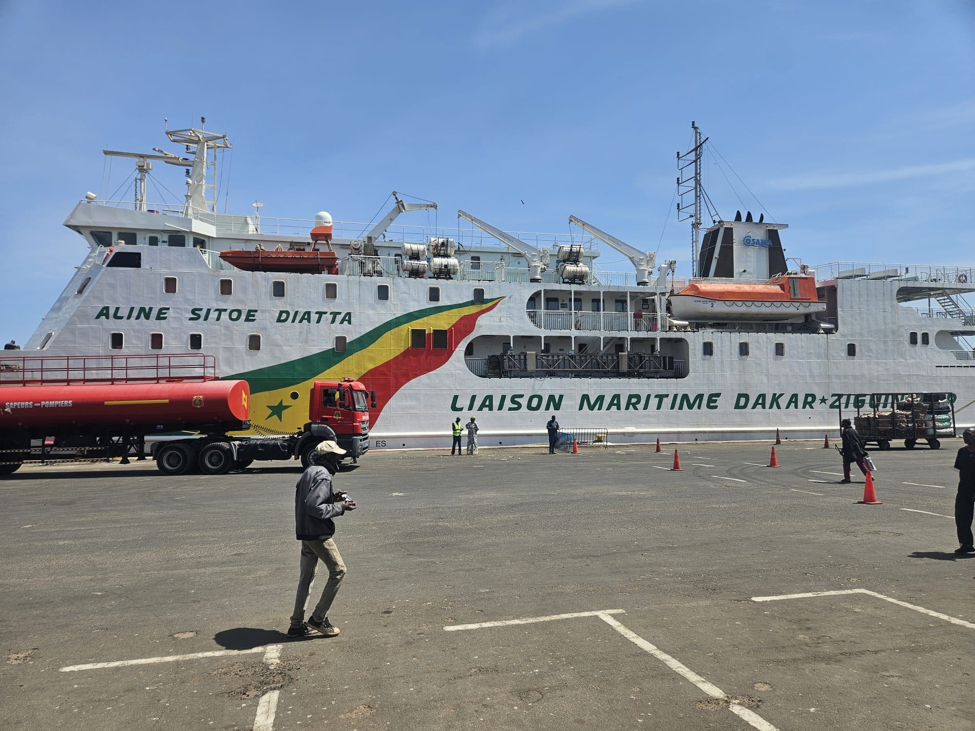 Een grote veerboot genaamd Aline Sitoe Diatta met de kleuren van de Senegalese vlag op de zijkant is aangemeerd in een haven. Vlakbij staan een rode tankwagen en een aantal mensen op de stoep onder een strakblauwe hemel.