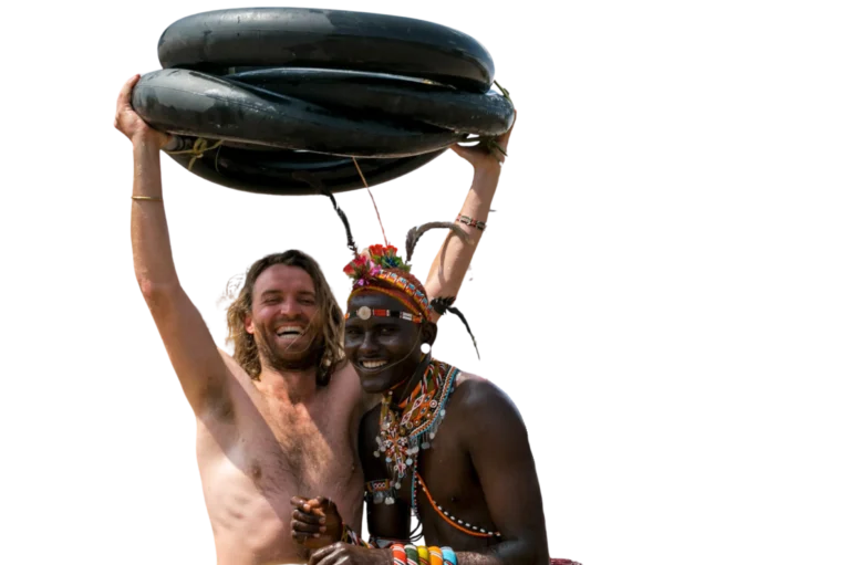 Twee lachende mannen, één zonder shirt en één in traditionele kralenkleding, staan bij elkaar. De shirtloze man tilt een aantal zwarte binnenbanden boven zijn hoofd terwijl ze er allebei blij en feestelijk uitzien.