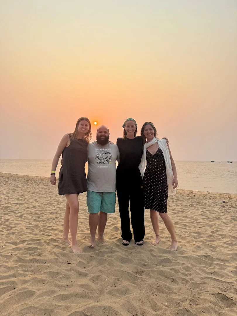 Vier mensen staan op blote voeten op een zandstrand bij zonsondergang en glimlachen naar de camera. De zon staat laag boven de kalme oceaan en de lucht is zacht oranje en roze. Twee vrouwen dragen jurken, één draagt een zwarte broek en één man draagt een korte broek.