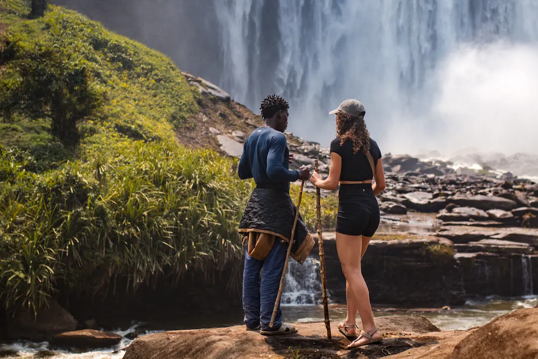 Twee mensen staan op rotsen bij een waterval en houden wandelstokken vast. De een draagt een hoed, zwart shirt en korte broek, terwijl de ander een blauw shirt met lange mouwen en een donkere broek draagt. Mist en groen omringen hen.