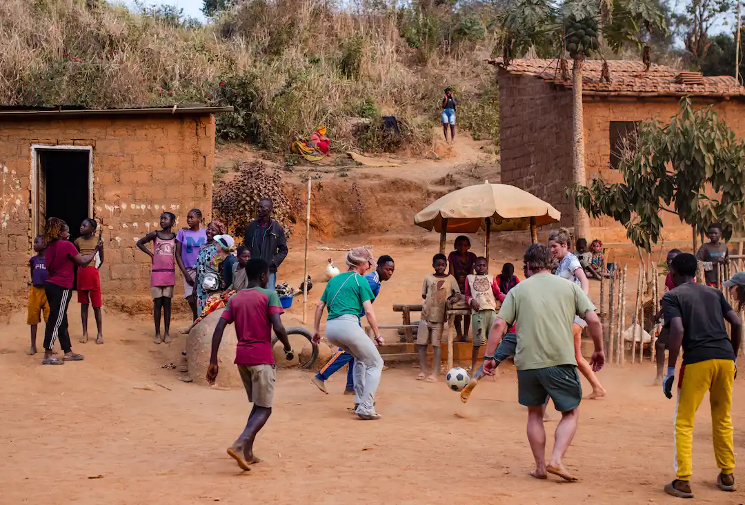 Een groep volwassenen en kinderen speelt voetbal op een stoffig veld in een plattelandsdorpje, met eenvoudige stenen huizen en omstanders die het spel gadeslaan. Sommige kinderen staan bij gebouwen, terwijl anderen van een afstandje toekijken.