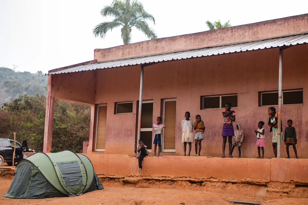 Een groep kinderen staat buiten een verweerd, perzikkleurig gebouw met een tinnen dak. Voor hen staat een groene kampeertent op de onverharde grond. Op de achtergrond zijn palmbomen en heuvels zichtbaar.