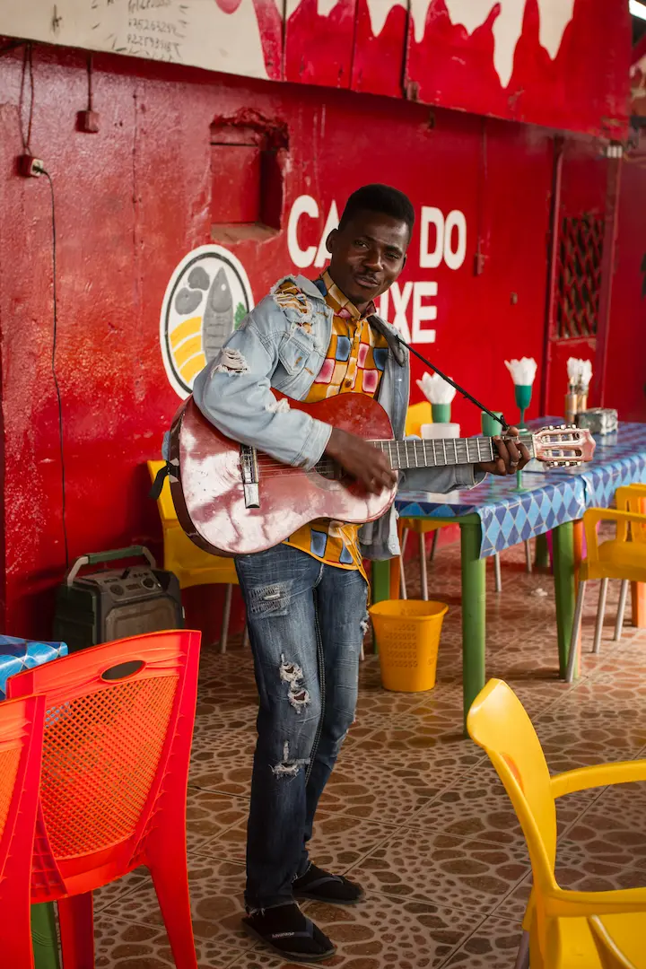 Een man in een spijkerjasje en gescheurde spijkerbroek speelt op een akoestische gitaar in een kleurrijk café met rode muren, gele en oranje stoelen en tafelkleden met blauwe patronen. Hij glimlacht en staat bij een tafel.