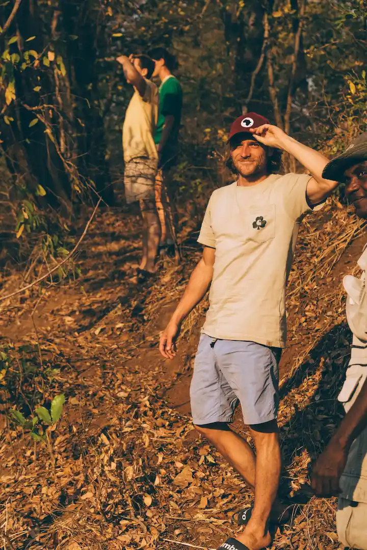 Tijdens een rondreis in Ghana staan mensen op een droge, met bladeren bedekte helling omringd door bomen. De voorste persoon draagt een licht T-shirt, een blauwe korte broek en een rode pet en lacht naar de camera. De anderen kijken in de verte.