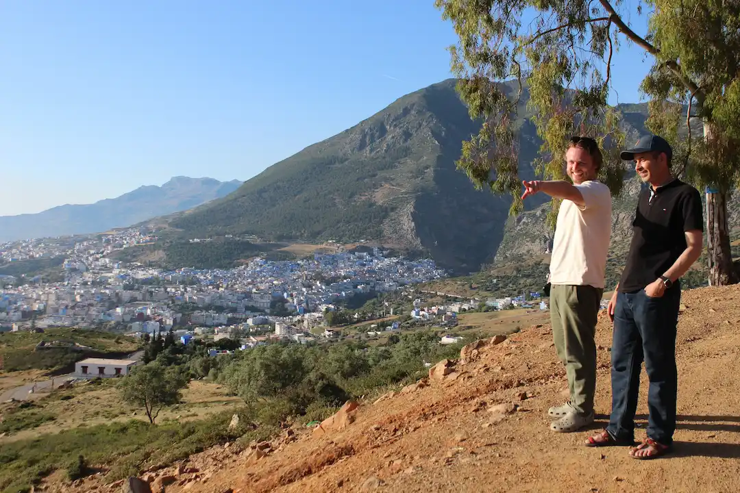Twee mannen staan op een heuvel; één van hen wijst naar een stadje in de verte, dat aan de voet van een grote groene berg ligt onder een helderblauwe hemel. Op de voorgrond staan bomen en rotsachtige grond.
