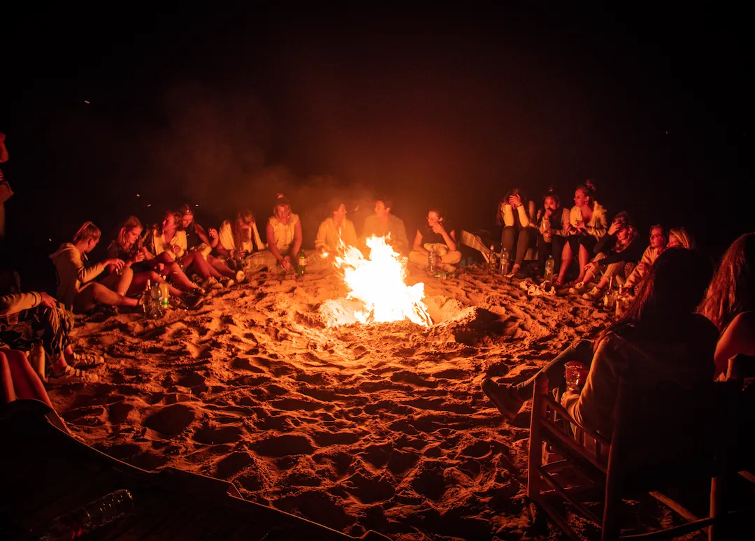 Een groep mensen zit 's nachts in een cirkel op zandgrond rond een groot kampvuur, verlicht door de gloed van het vuur, met duisternis om hen heen.