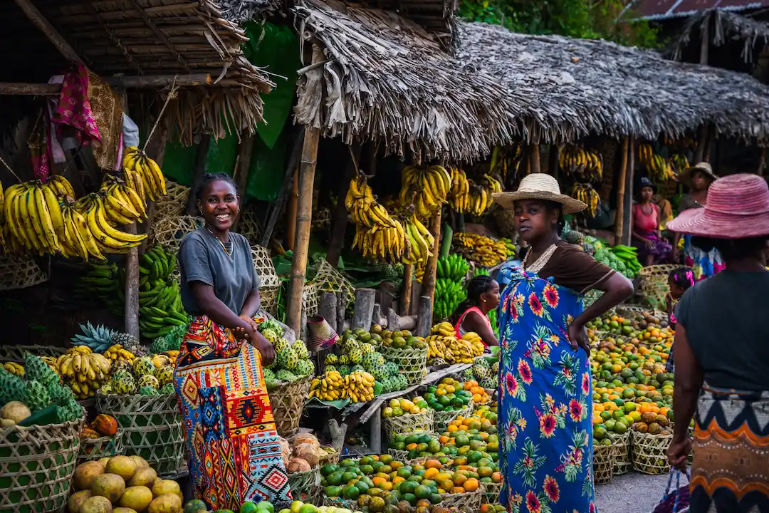 Vrouwen in kleurrijke jurken staan glimlachend op een levendige openluchtmarkt, omringd door manden met fruit en groenten, waaronder bananen, mango’s en papaja’s, onder kraampjes met rieten daken.