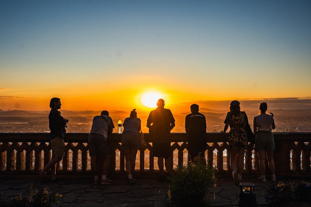 Zeven mensen staan tegen een stenen balustrade geleund en kijken naar een feloranje zonsondergang boven het stadslandschap beneden, met een heldere hemel en wat groen op de voorgrond.