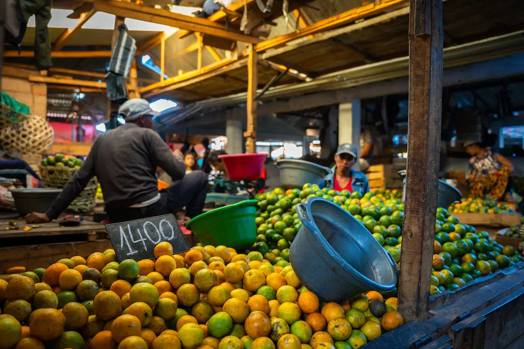 Op een marktkraam liggen stapels groene en oranje citrusvruchten, met daarbovenop een metalen bak. Op een bord staat 1400. Op de achtergrond zijn onder een houten dak verkopers en andere kraampjes te zien.