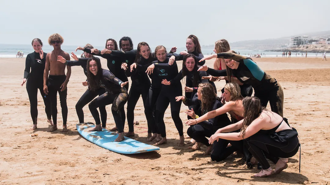 Een groep mensen in wetsuits poseert speels rond een blauwe surfplank op een zandstrand, glimlacht en maakt leuke gebaren, met de oceaan en strandgangers in de verte op de achtergrond.