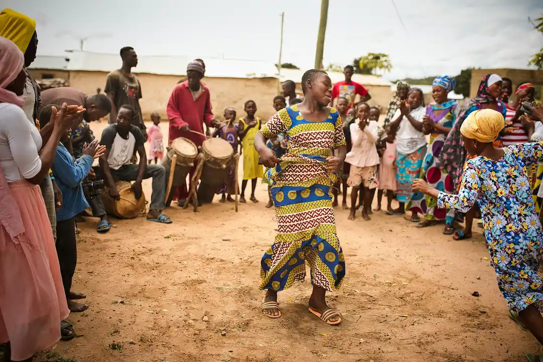 Tijdens een rondreis in Ghana verzamelt een groep mensen zich buiten, klapt en juicht terwijl twee vrouwen in het midden dansen. Drummers spelen op de achtergrond en kinderen en volwassenen kijken toe en vieren samen feest.