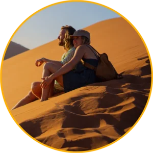 Een man en een vrouw zitten dicht tegen elkaar aan op een zonovergoten zandduin, glimlachen en genieten van het woestijnlandschap. De vrouw draagt een hoed en heeft een rugzak; beiden zien er ontspannen uit. De afbeelding wordt omlijst door een cirkelvormige gele rand.