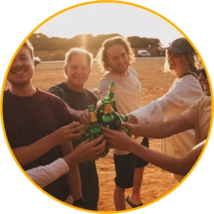 Een groep vrienden staat buiten bij zonsondergang bierflesjes te klinken en te glimlachen, omlijst door een grote gele cirkel.