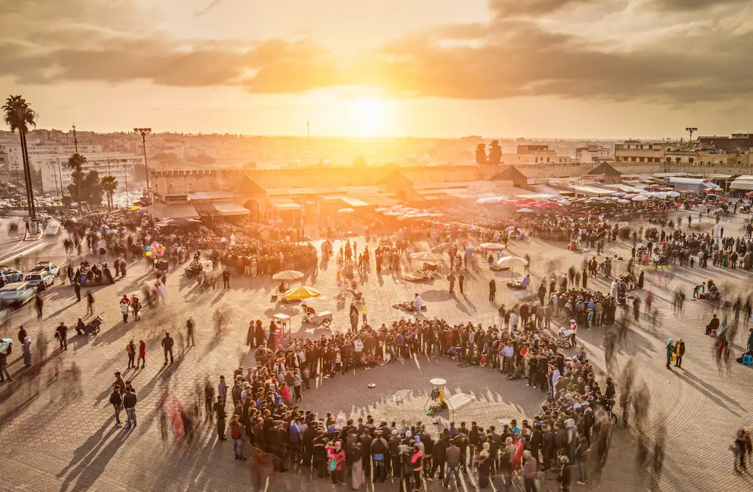Tijdens zonsondergang verzamelt zich een grote menigte in een kring op een open plein; op de achtergrond zijn vage bewegingen te zien die wijzen op levendige bedrijvigheid en marktkraampjes.
