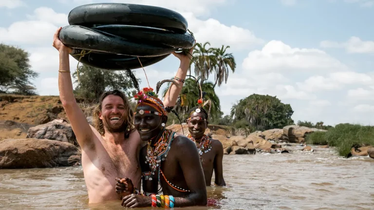 Drie glimlachende mensen staan in een rivier; één van hen houdt zwembanden boven zijn hoofd. Twee van hen dragen kleurrijke sieraden van kralen en hoofdtooien, terwijl de derde geen shirt aan heeft. Op de achtergrond zijn bomen, rotsen en een blauwe lucht te zien.