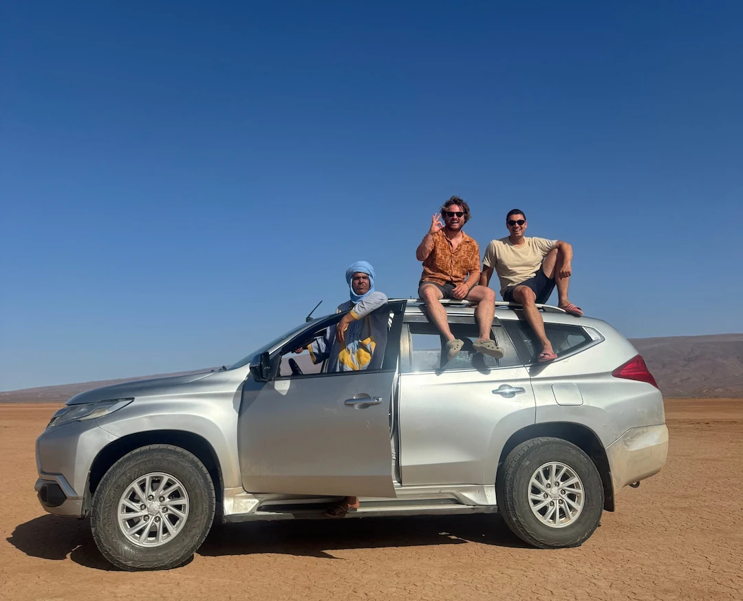 Drie mensen zitten op en in een zilverkleurige SUV die geparkeerd staat in een woestijnlandschap met een helderblauwe lucht en bergen in de verte. De autodeur staat open en de groep ziet er ontspannen en opgewekt uit.