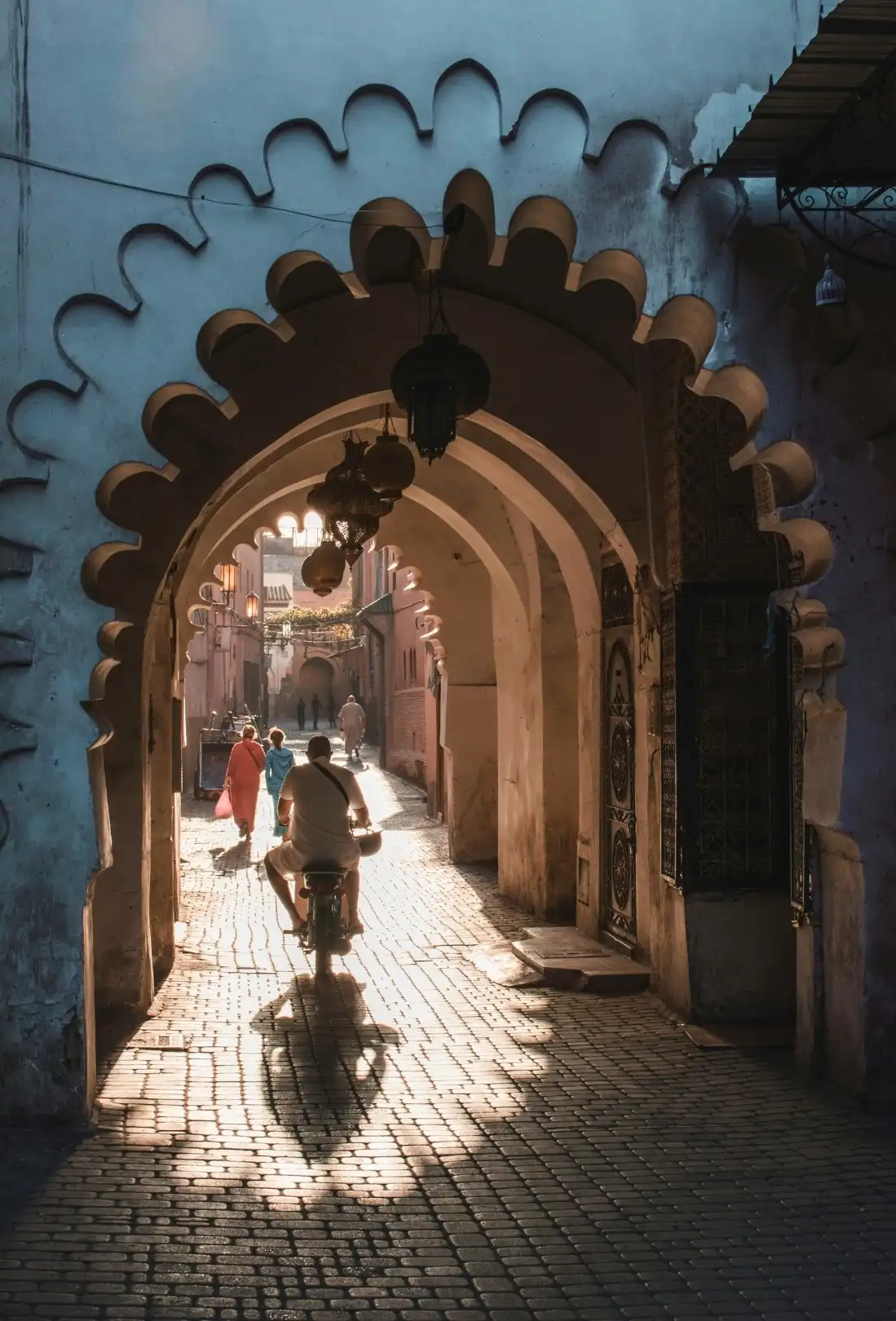 Iemand fietst door een sierlijke boogpoort in een straat met kasseien, terwijl het zonlicht schaduwen werpt. In de verte lopen verschillende mensen, en boven hun hoofden hangen sierlijke lantaarns.
