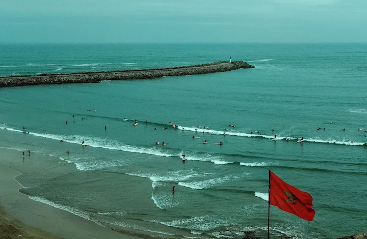Op een zandstrand wappert een rode vlag met een groene ster, terwijl talrijke surfers en zwemmers onder een bewolkte hemel genieten van de turkooizen golven bij een lange stenen pier die zich uitstrekt in de oceaan.