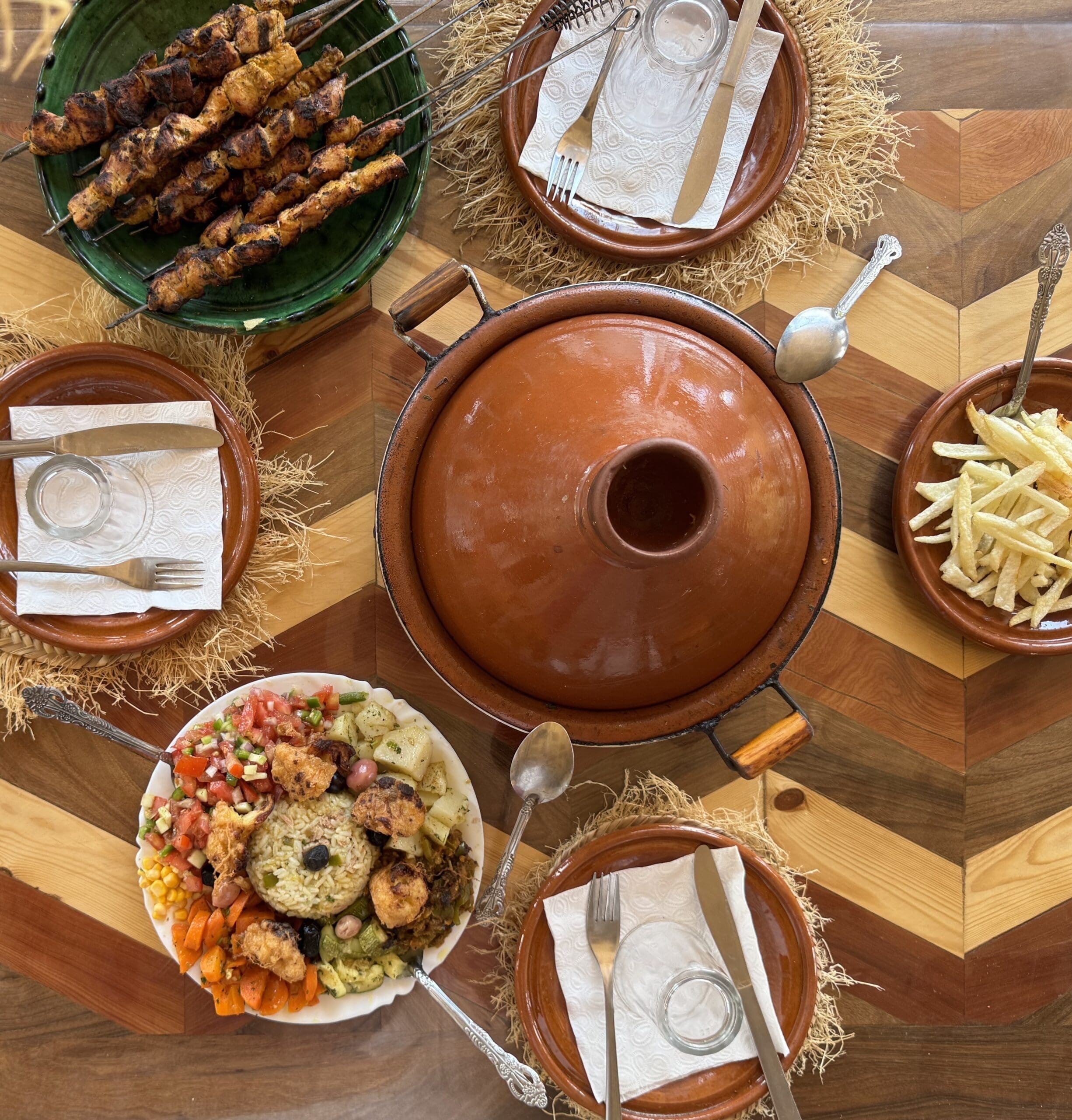 Een tafel waarop in het midden een grote aardewerken pot staat, met daarnaast borden met gegrilde spiesjes, frietjes, een kleurrijk gerecht met rijst en groenten, en een gedekte tafel met borden, glazen en bestek op een houten tafel met chevronpatroon.