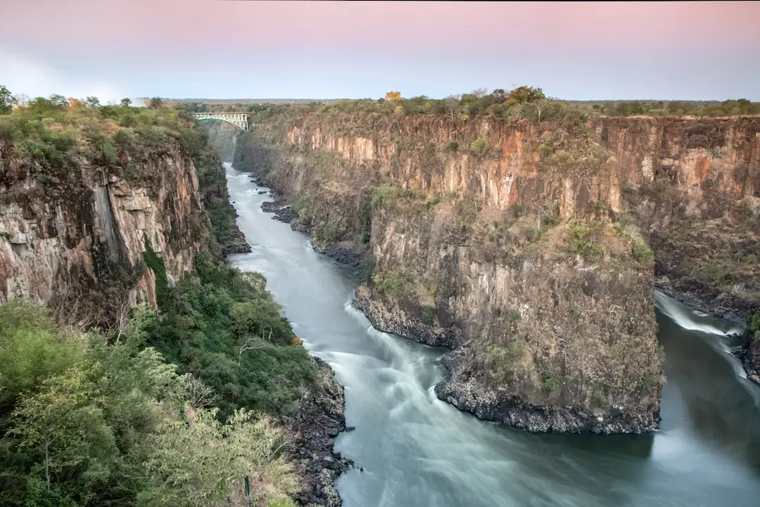 Een brede rivier stroomt tussen steile, rotsachtige kliffen met schaarse begroeiing; in de verte overspant een brug de kloof onder een pastelkleurige hemel.