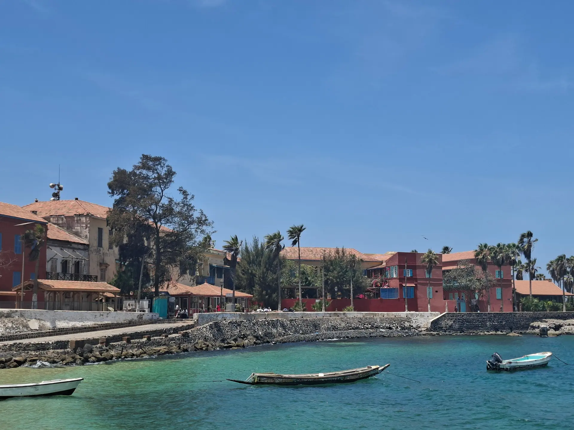 Drie kleine houten bootjes dobberen op helder turquoise water vlakbij een rotsachtige kustlijn, met op de achtergrond verschillende gebouwen met rode daken, palmbomen en een stralend blauwe lucht.