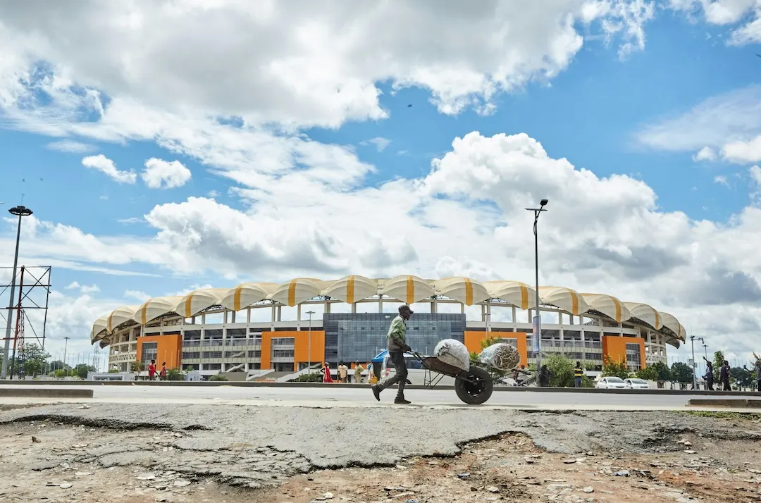 Een persoon duwt een kruiwagen vol met grote zakken langs een modern stadion, onder een blauwe hemel met hier en daar wat wolken.