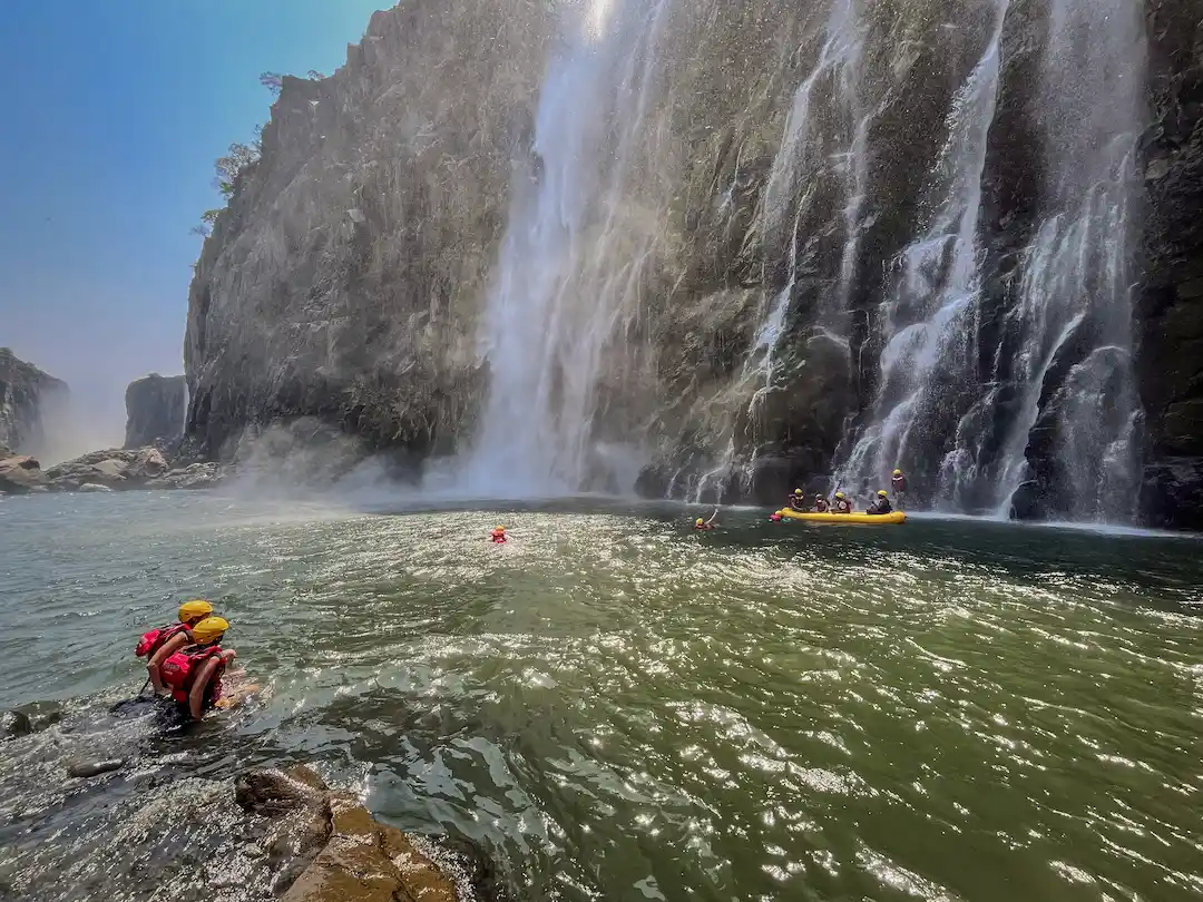 Mensen zwemmen en varen op vlotten in de buurt van een grote waterval die van een rotsachtige klif naar beneden stort. Er stijgt mist op uit het water, en boven de waterval is weelderig groen te zien onder een stralend blauwe hemel.