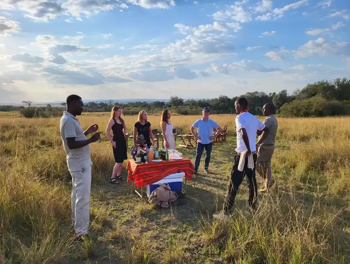 Zes mensen staan rond een tafel met drankjes op een oranje gestreept tafelkleed in een grasveld onder een blauwe hemel met wolken; op de achtergrond zijn nog meer tafels en stoelen te zien.