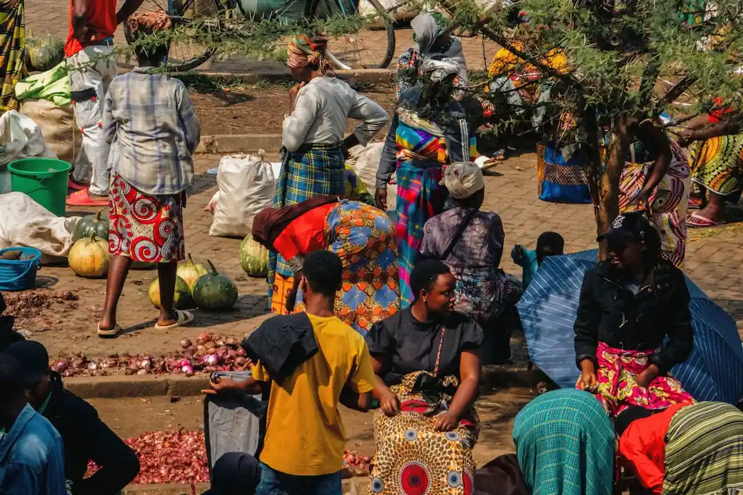 Mensen verzamelen zich op een openluchtmarkt, waar ze goederen zoals uien en watermeloenen kopen en verkopen. Velen dragen kleding met kleurrijke patronen. Het is er een levendig tafereel: sommige kopers staan, terwijl anderen tussen de manden en tassen zitten.