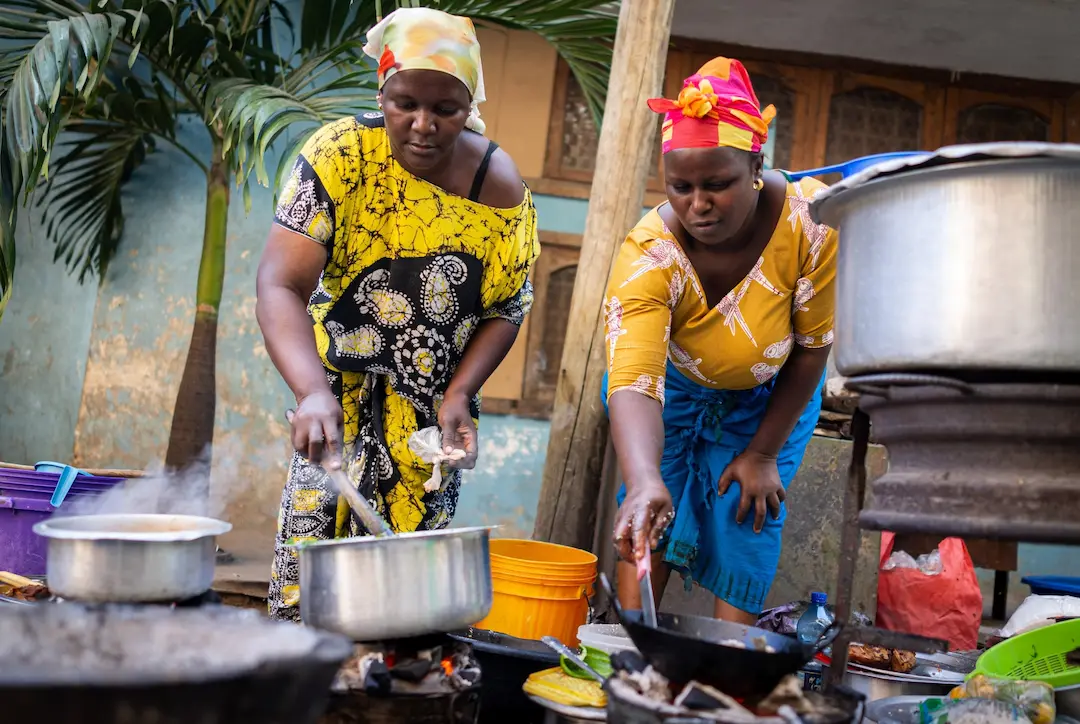 Twee vrouwen in kleurrijke kleding bereiden buiten eten, terwijl ze in potten boven open vuur roeren. Ze worden omringd door grote metalen potten, keukengerei en diverse bakjes. Op de achtergrond staan een palmboom en een gebouw.