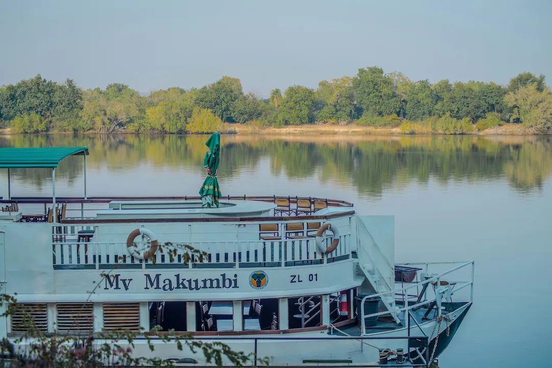 Een witte boot met de naam Mv Makumbi ligt aangemeerd in rustig water, met groene bomen en een heldere hemel op de achtergrond.