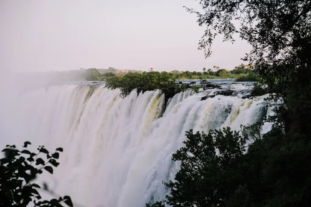 Een grote waterval stort zich over een rotsachtige rand, omringd door weelderige groene bomen en begroeiing onder een bleke hemel. Er stijgt mist op uit het krachtig neerstortende water.