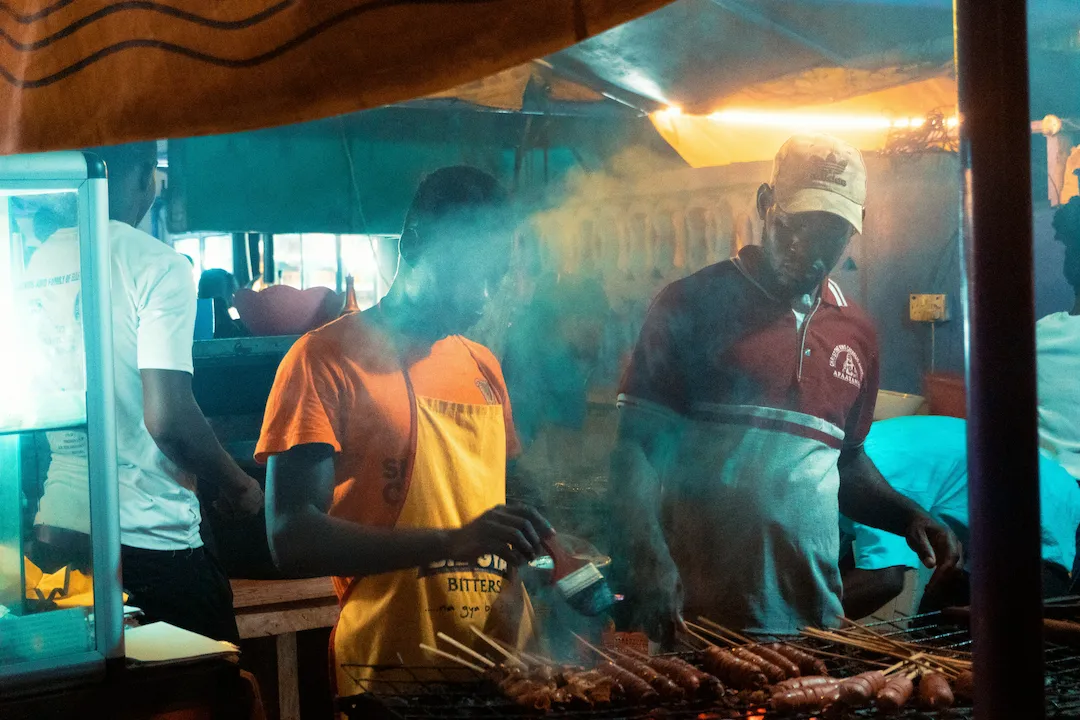 Twee mannen grillen vlees aan spiesen bij een rokerig eetkraampje op een avondmarkt in de open lucht; de ene draagt een oranje shirt en de andere een rode polo en een pet, onder kleurrijke verlichting.