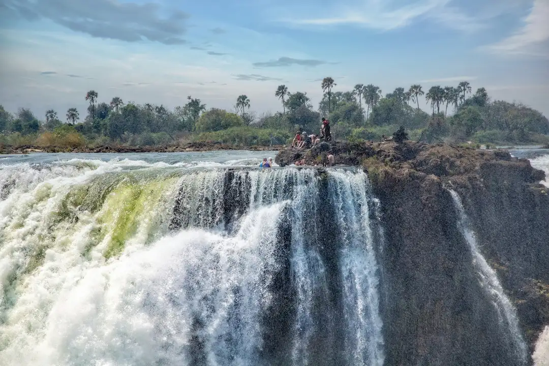 Een aantal mensen staat op rotsen aan de rand van een grote waterval, waar het water over de rand naar beneden stort. Op de achtergrond zijn bomen en groen te zien onder een blauwe lucht met hier en daar wat wolken.